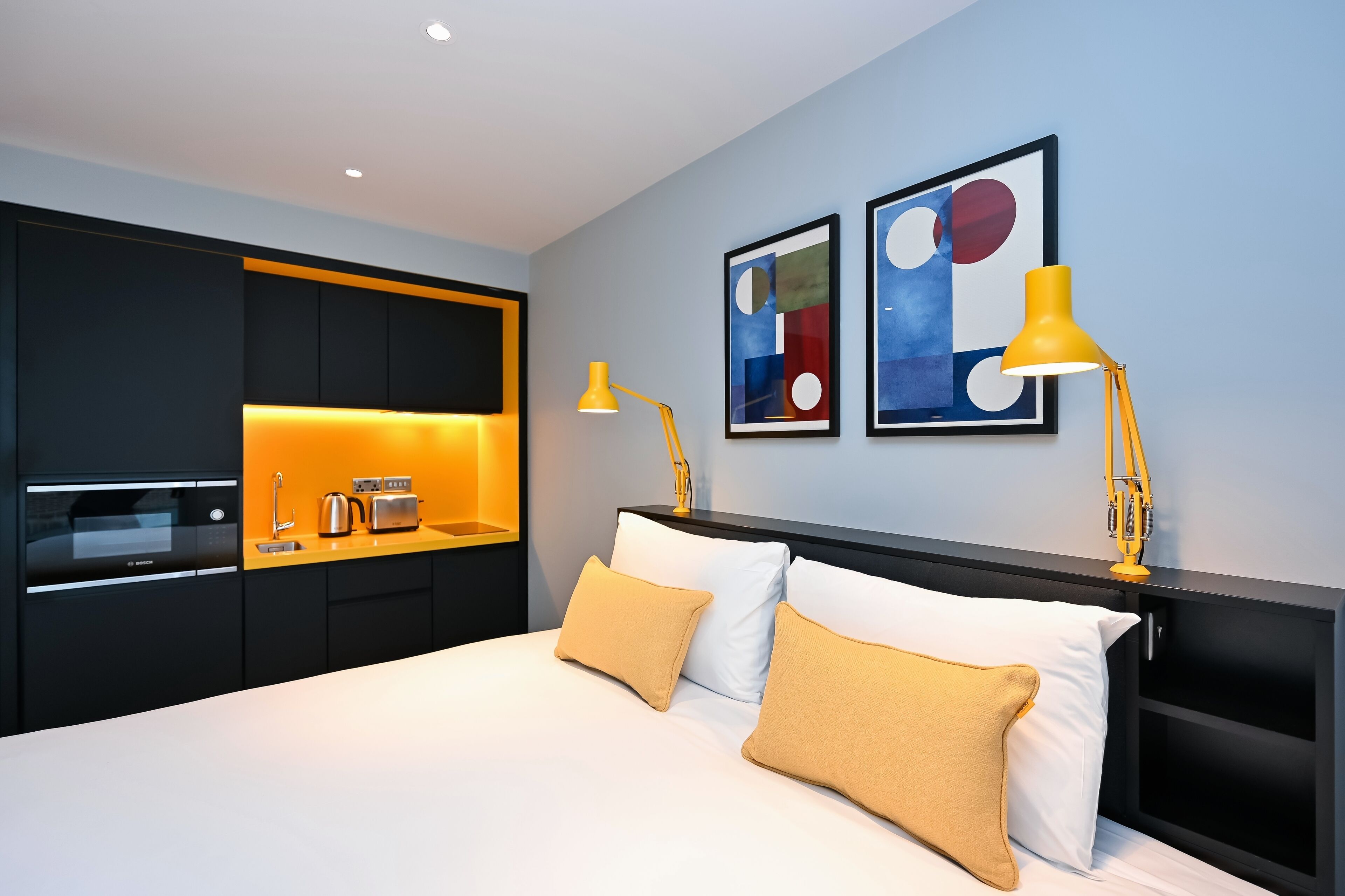 A modern hotel room with a bed, two bedside lamps, a kitchen area, and abstract wall art.