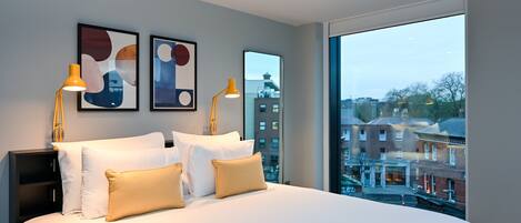 A modern hotel room with a large bed, two bedside lamps, and a city view through a large window.