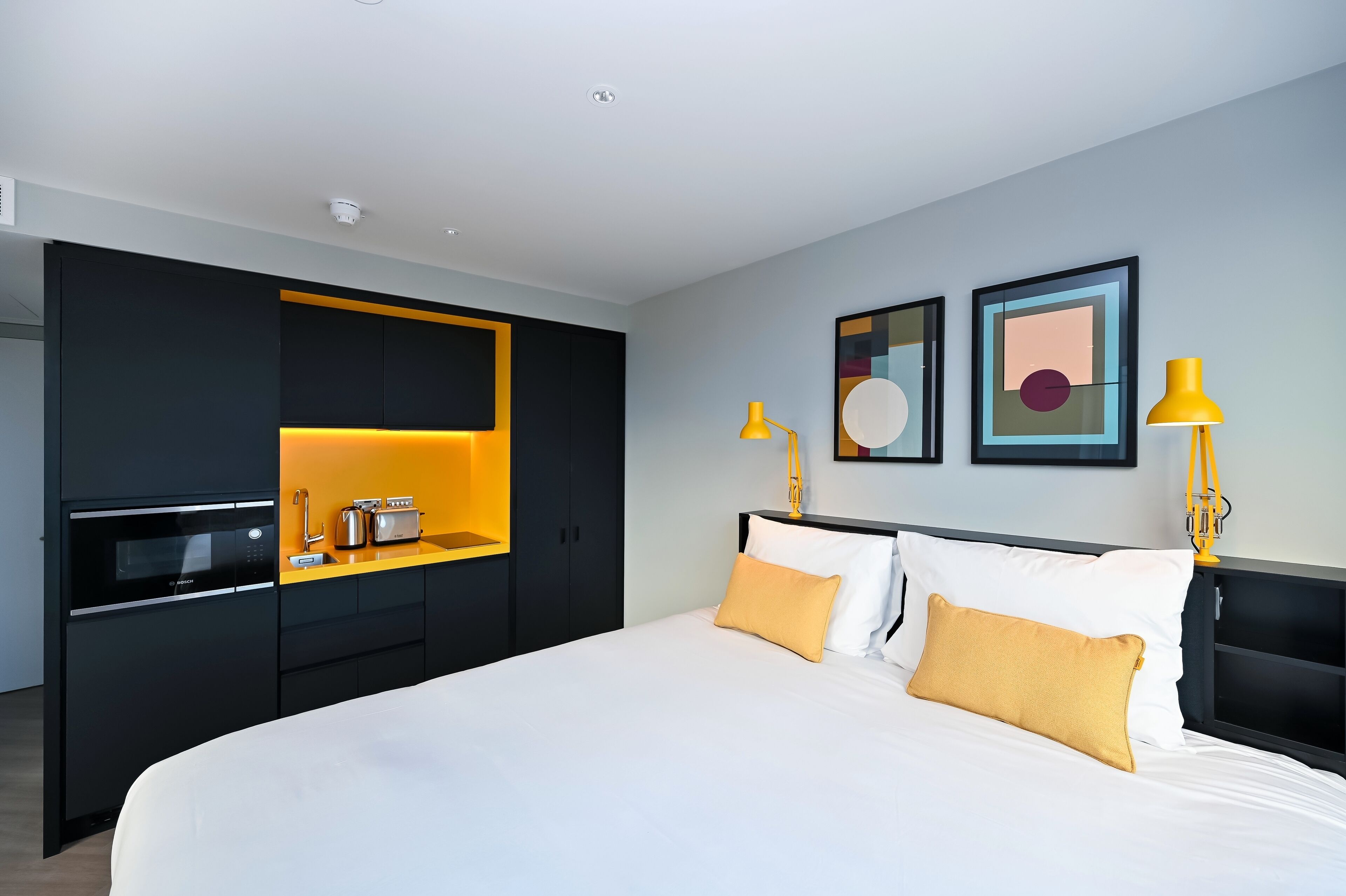 A modern hotel room with a large bed, a built-in kitchen with a microwave, toaster, and sink, and two wall-mounted lamps.