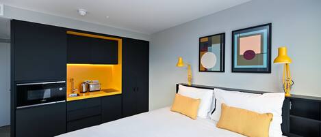 A modern hotel room with a large bed, a built-in kitchen with a microwave, toaster, and sink, and two wall-mounted lamps.