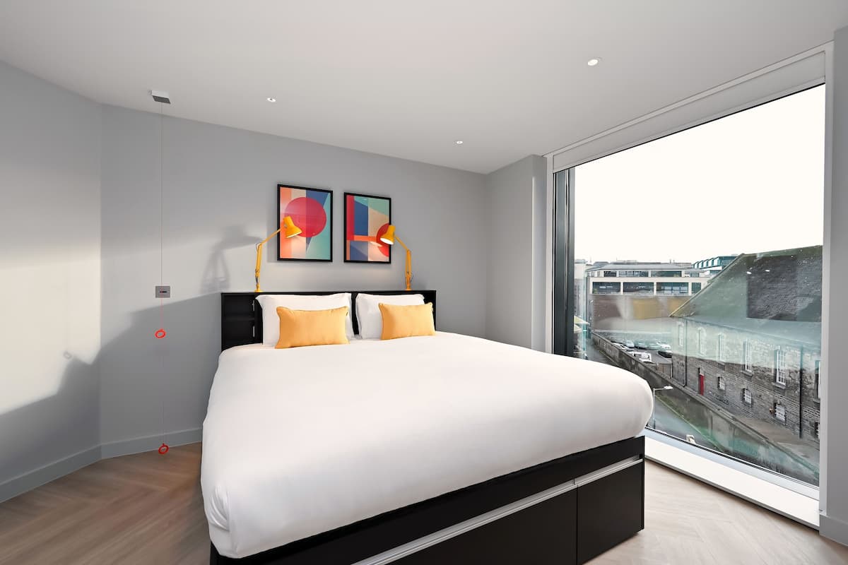Ein modernes Schlafzimmer mit einem großen Bett, Blick auf die Stadt durch ein großes Fenster und zwei abstrakte Gemälde an der Wand.