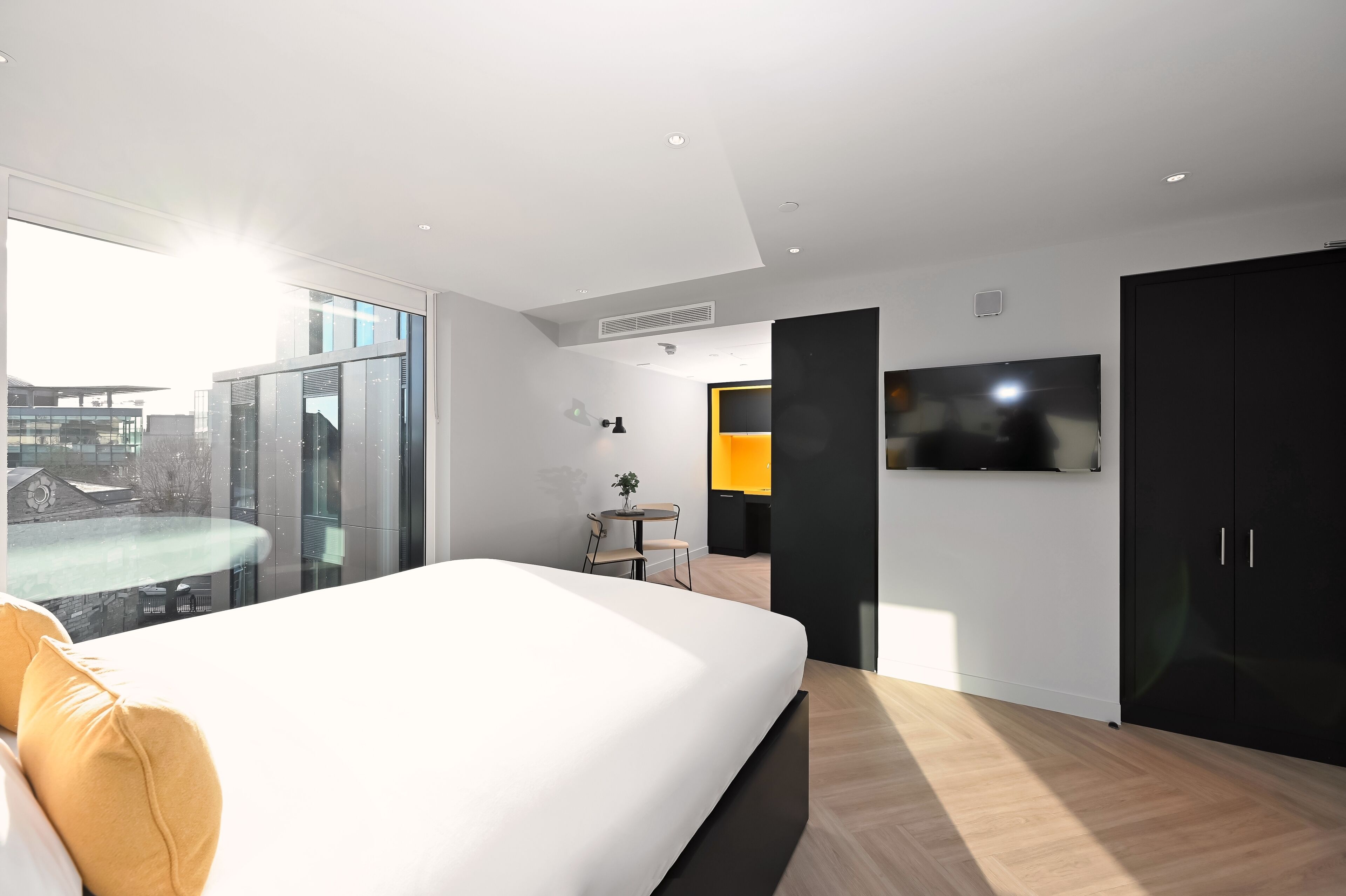 Una habitación de hotel moderna con una cama grande, un televisor montado en la pared y vistas a la ciudad a través de amplios ventanales.