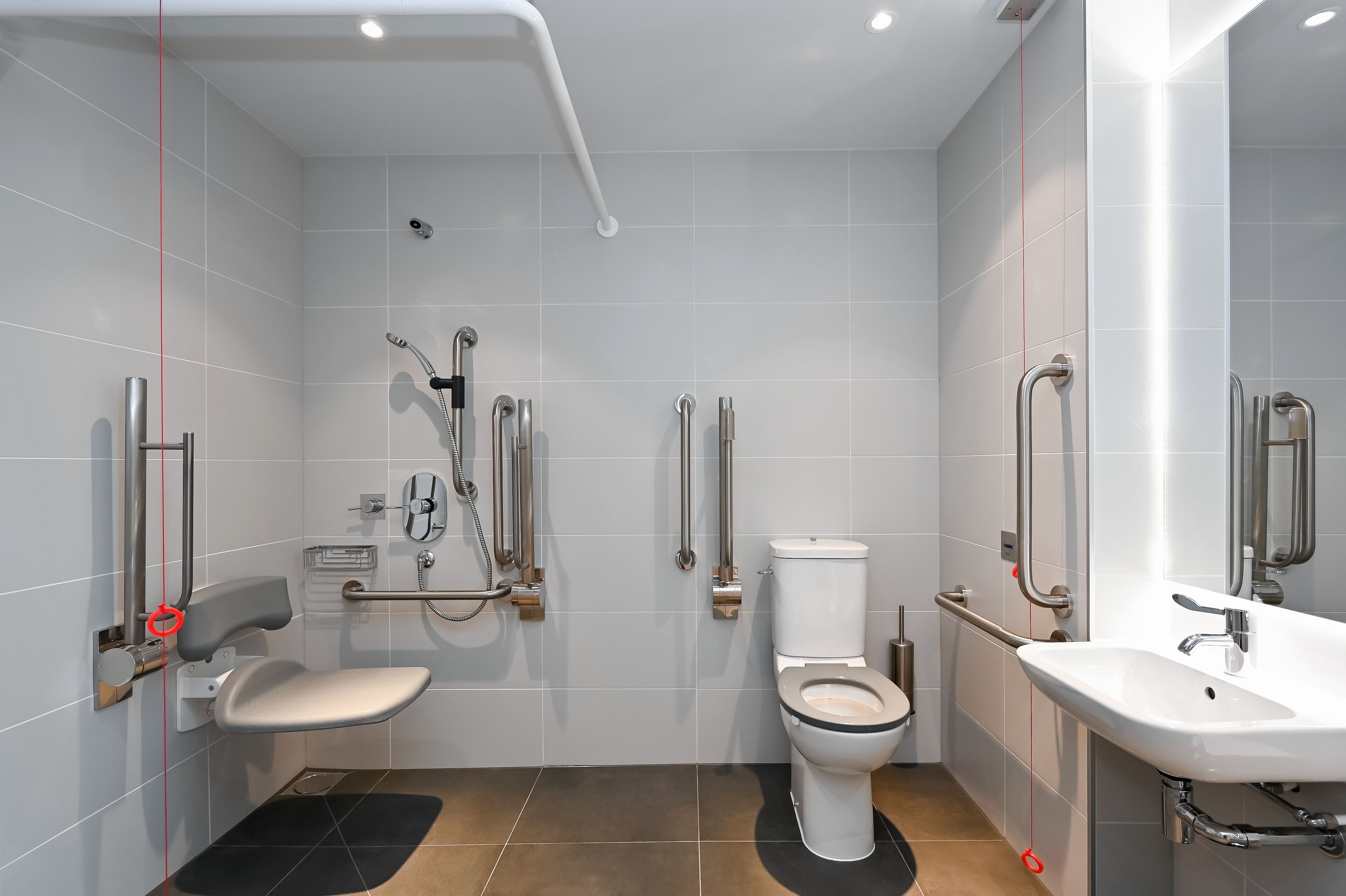 A modern bathroom with a toilet, bidet, and sink, equipped with accessibility features like grab bars and a shower chair.