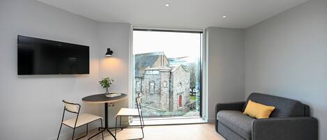 A modern living room with a grey sofa, a small round table, and two chairs. A large window offers a view of a stone building.