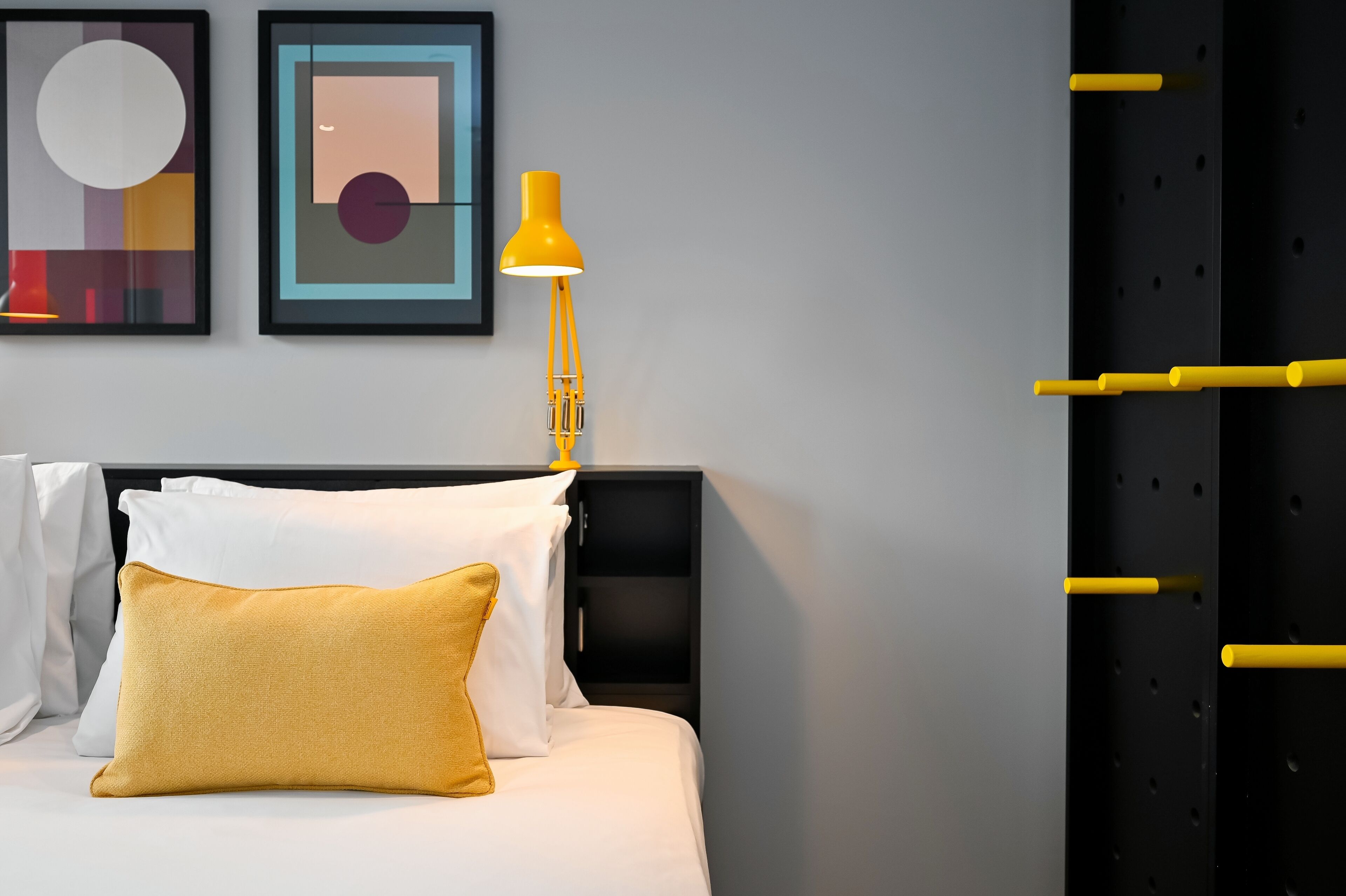 A neatly made bed with white and mustard pillows, a black headboard, and a yellow lamp.