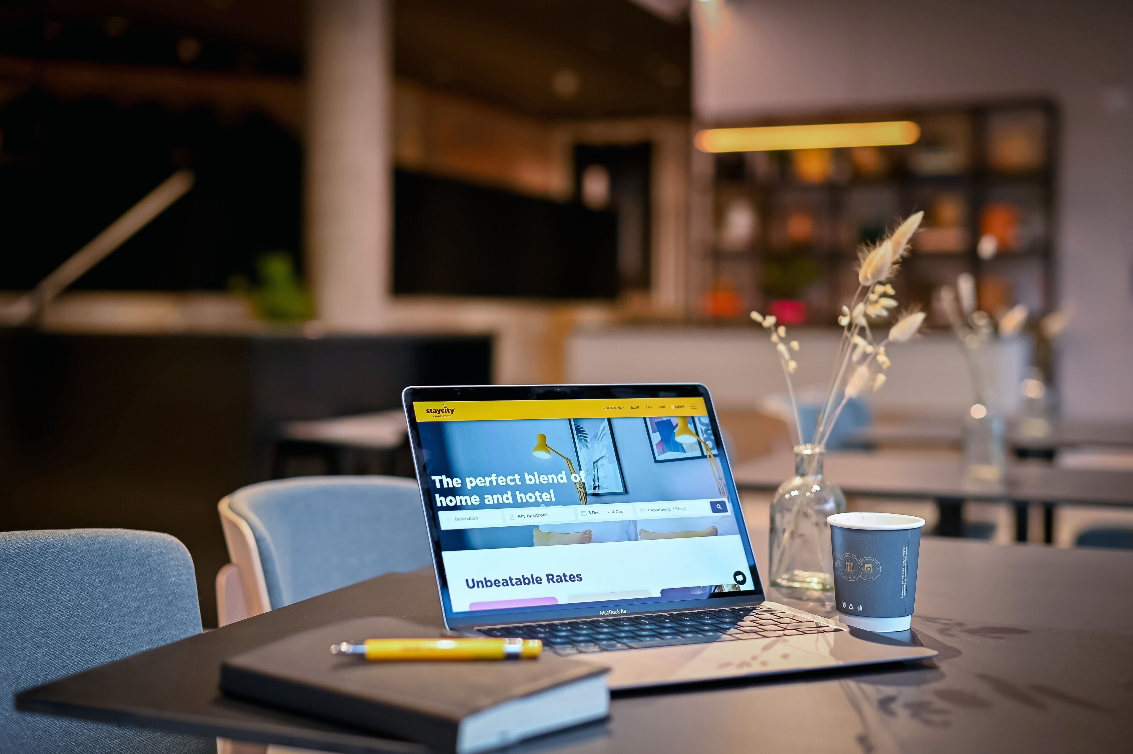 A laptop displaying a hotel booking website, a coffee cup, a notebook, and a pen on a table.