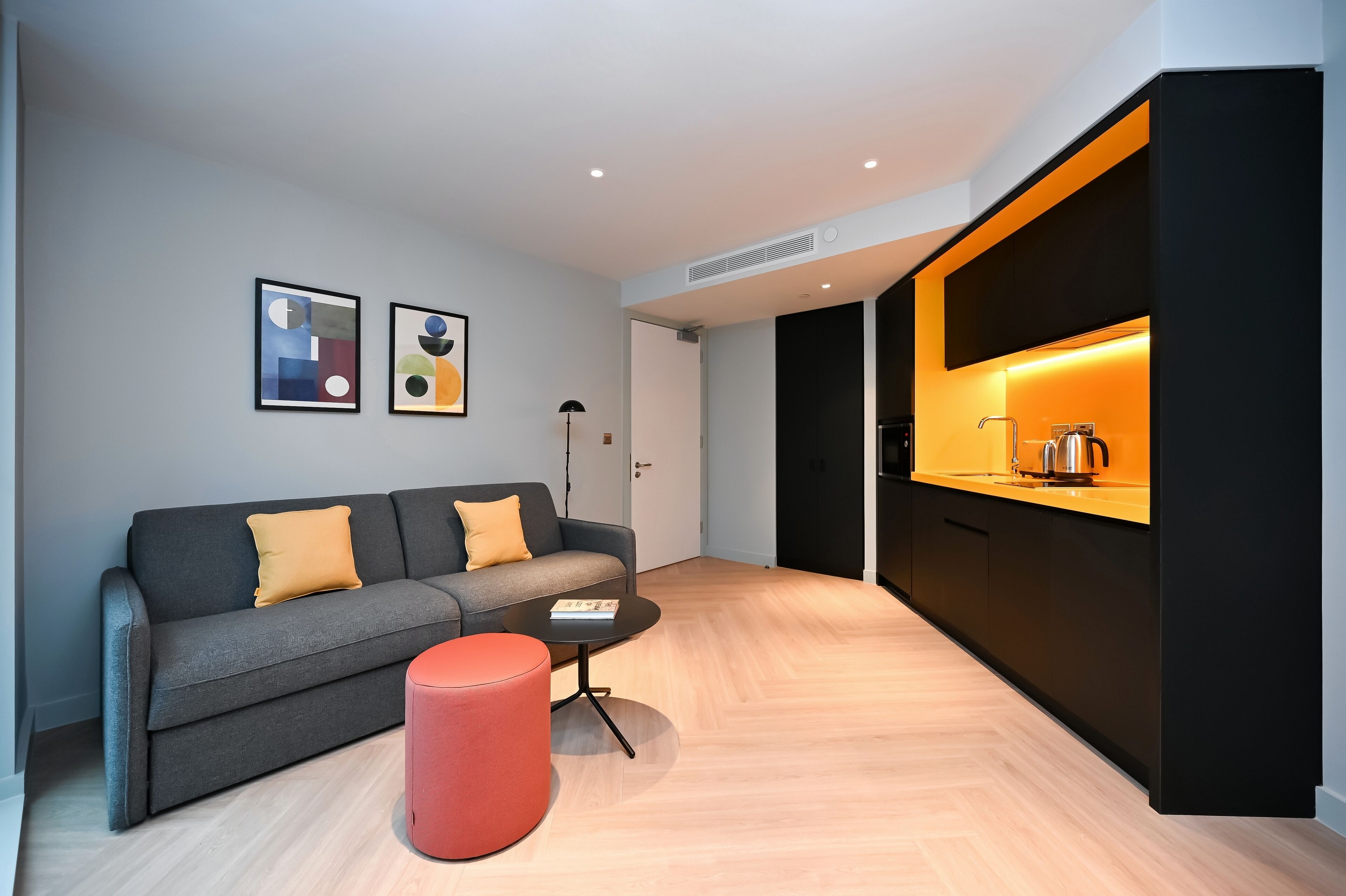 A modern living room with a grey sofa, red ottoman, and a coffee table. There are two framed artworks on the wall and a kitchen area with a black countertop and orange lighting.