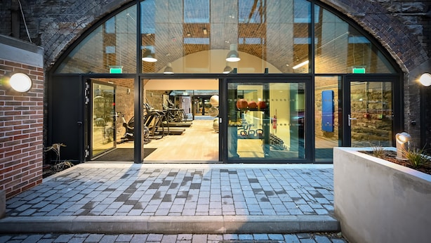 Un bâtiment moderne doté d’une grande entrée en verre en forme d’arc, d’une façade en briques et d’un chemin pavé menant à l’entrée.