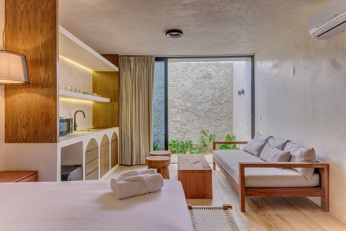 Quarto de hotel moderno com cama, sofá, pequena cozinha e varanda com plantas.