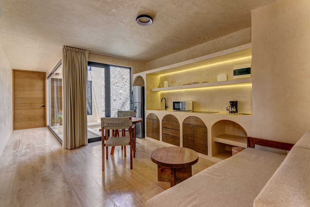 Uma sala de estar moderna com sofá, mesa redonda de madeira e área de jantar com cadeiras. Há também uma cozinha com micro-ondas e prateleiras.