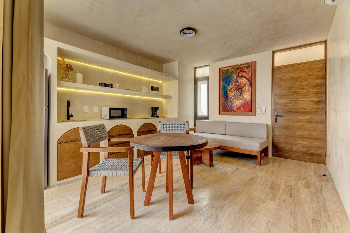 Uma sala de estar moderna com uma mesa de madeira redonda, duas cadeiras, um sofá e uma cozinha planejada, com micro-ondas e prateleiras.