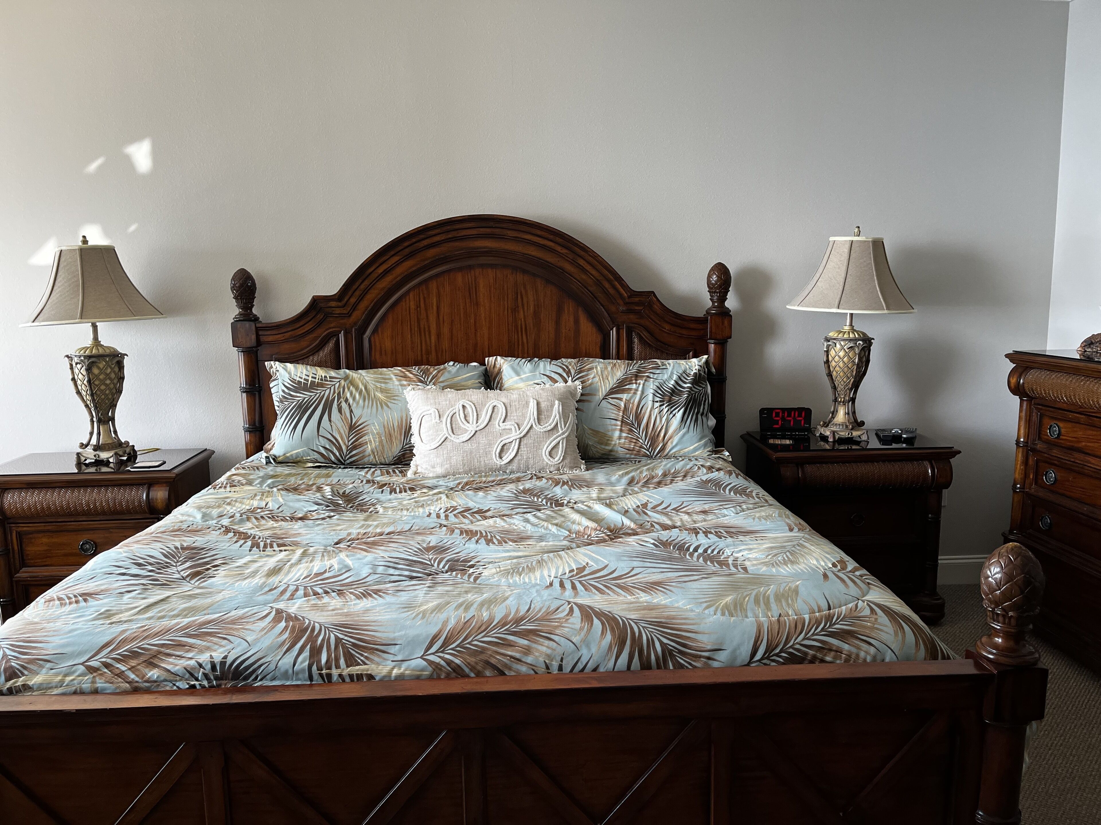 Un lit bien fait, recouvert d’une couvre-lit à motifs tropicaux, flanqué de deux tables de chevet en bois avec des lampes, d’une tête de lit en bois et d’une commode en bois.