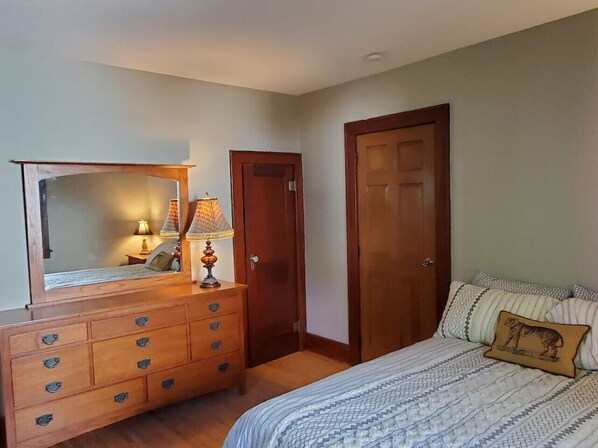 A bedroom with a wooden dresser, a mirror, a lamp, a bed with patterned bedding, and two doors.