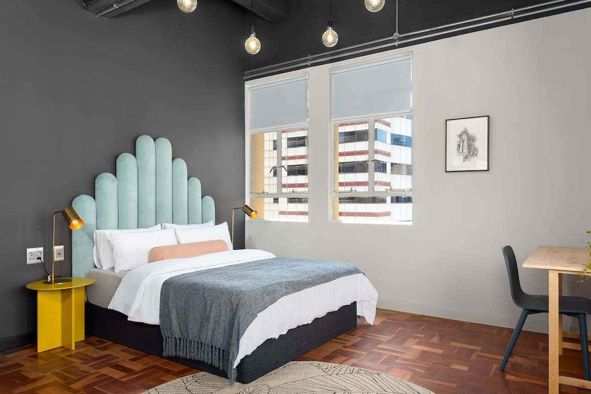 A modern bedroom with a tufted headboard, a bed with white and grey bedding, a yellow bedside table with a lamp, a wooden floor, and a window with a view of buildings.