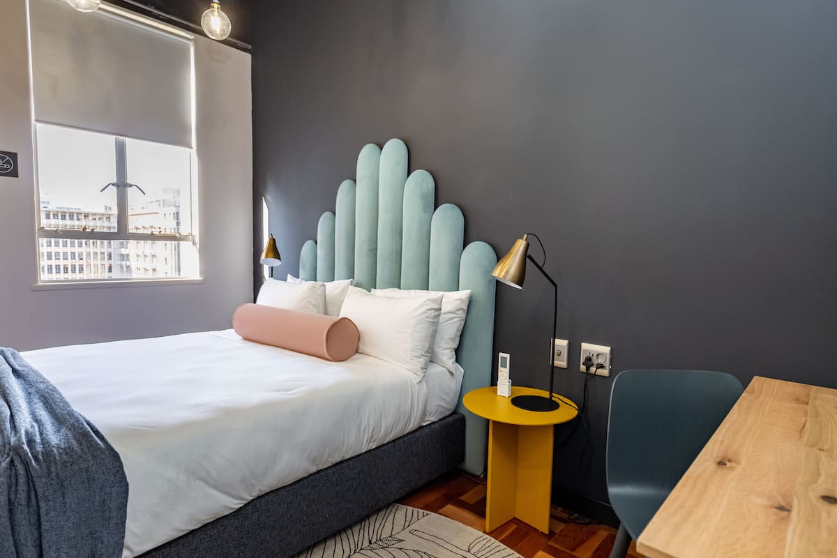 A modern bedroom with a bed, bedside table, lamp, chair, and a view of the city through a window.