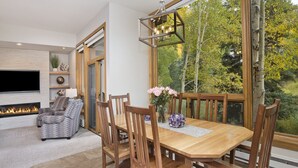 Un coin repas avec une table et des chaises en bois, une cheminée et une grande fenêtre donnant sur les arbres.