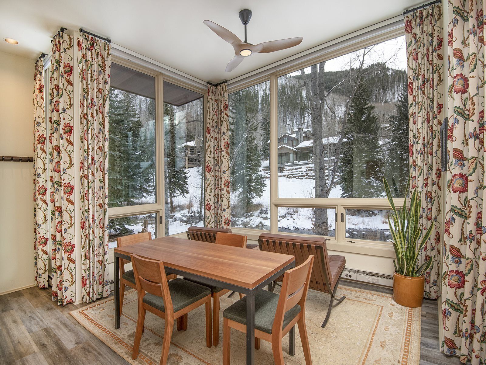 A dining area with a wooden table and chairs, large windows with floral curtains, a ceiling fan, and a view of a snowy forest.