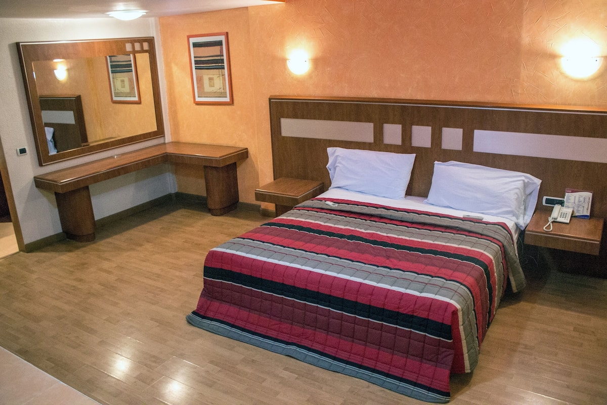A hotel room with a large bed, a bedside table, a nightstand, a mirror, and a wall-mounted light fixture.