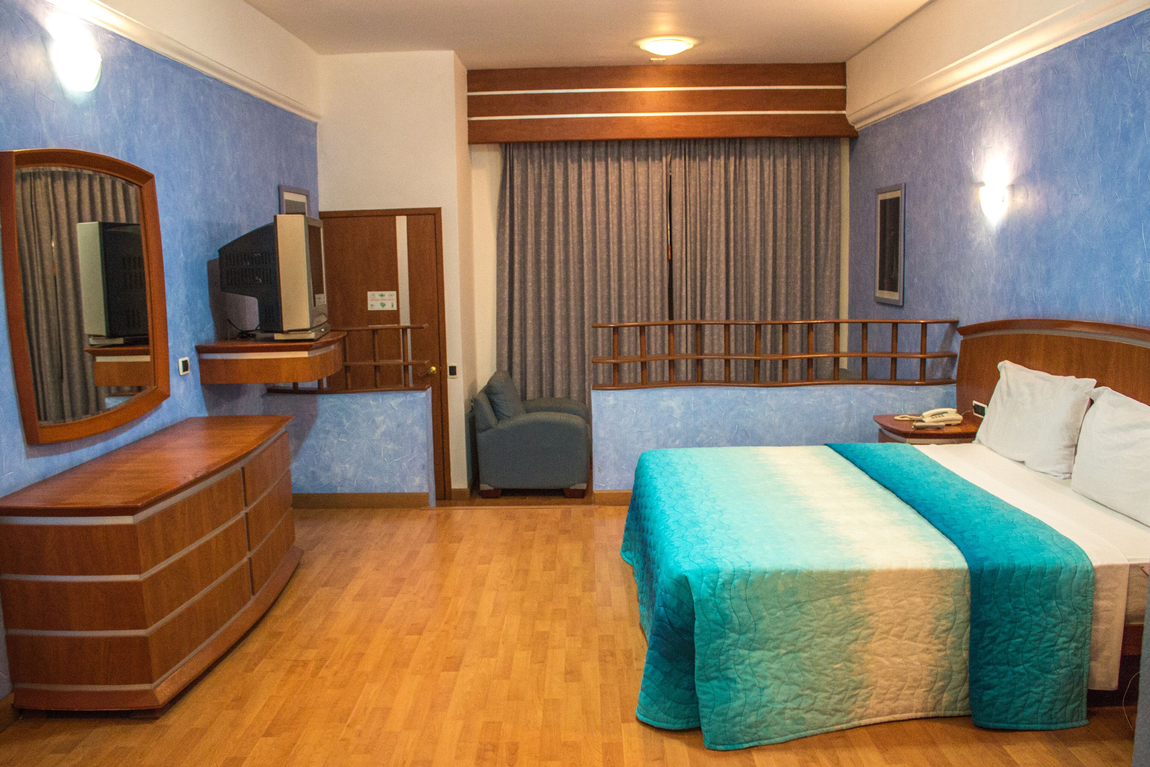 Hotellihuone, jossa on puinen lipasto, televisio, turkoosinsinisellä peitteellä varustettu sänky, yöpöytä ja tuoli.