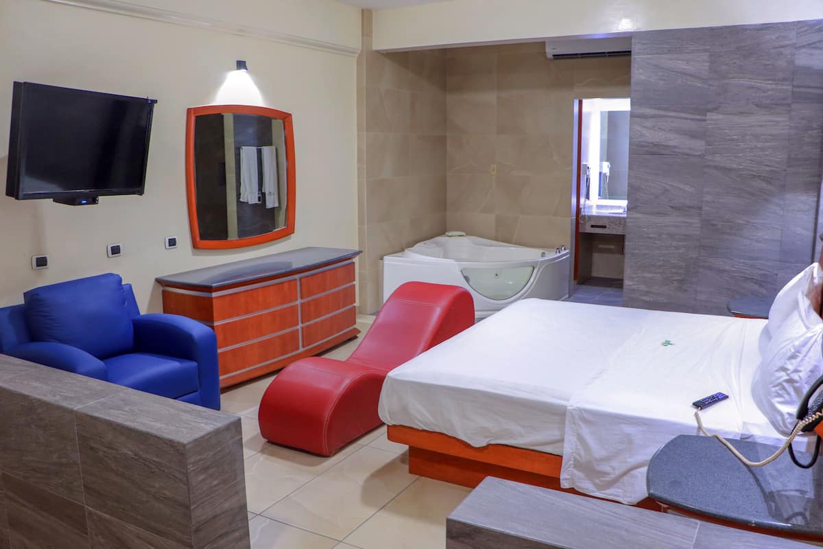 A hotel room with a bed, a red massage chair, a blue sofa, a wooden dresser, a TV, and a bathroom with a bathtub.