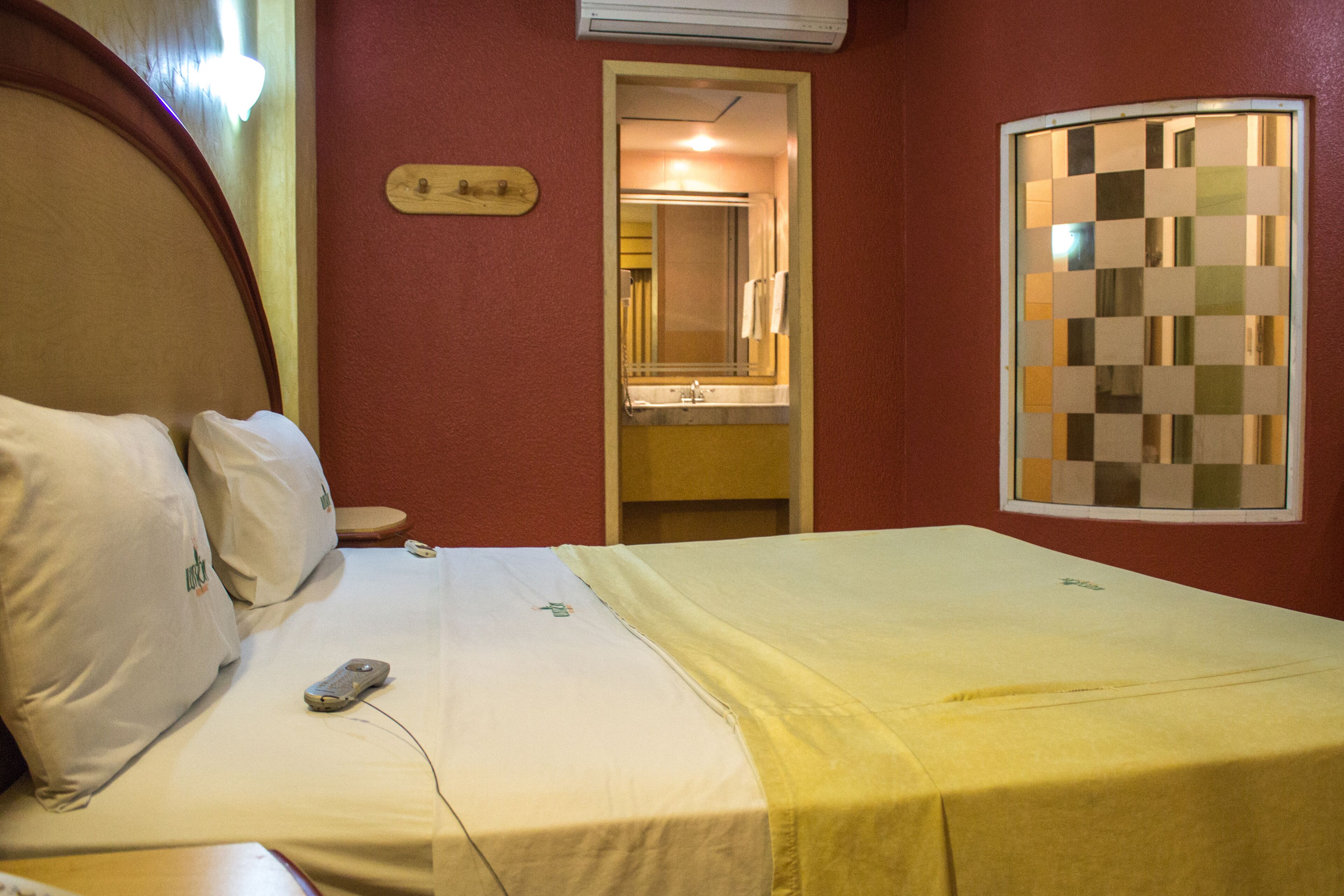 A hotel room with a bed, a bedside table, a remote control, a mirror, and a checkered window.