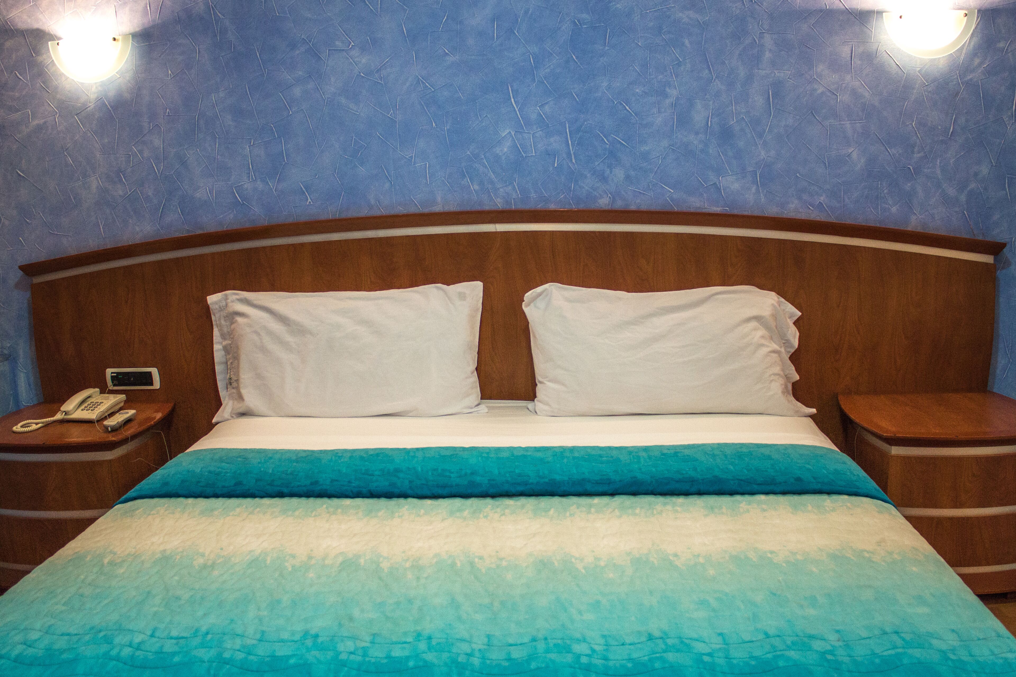 Hotelkamer met een groot bed, twee witte kussens, een houten hoofdbord, een nachtkastje met een telefoon en een blauwe muur.