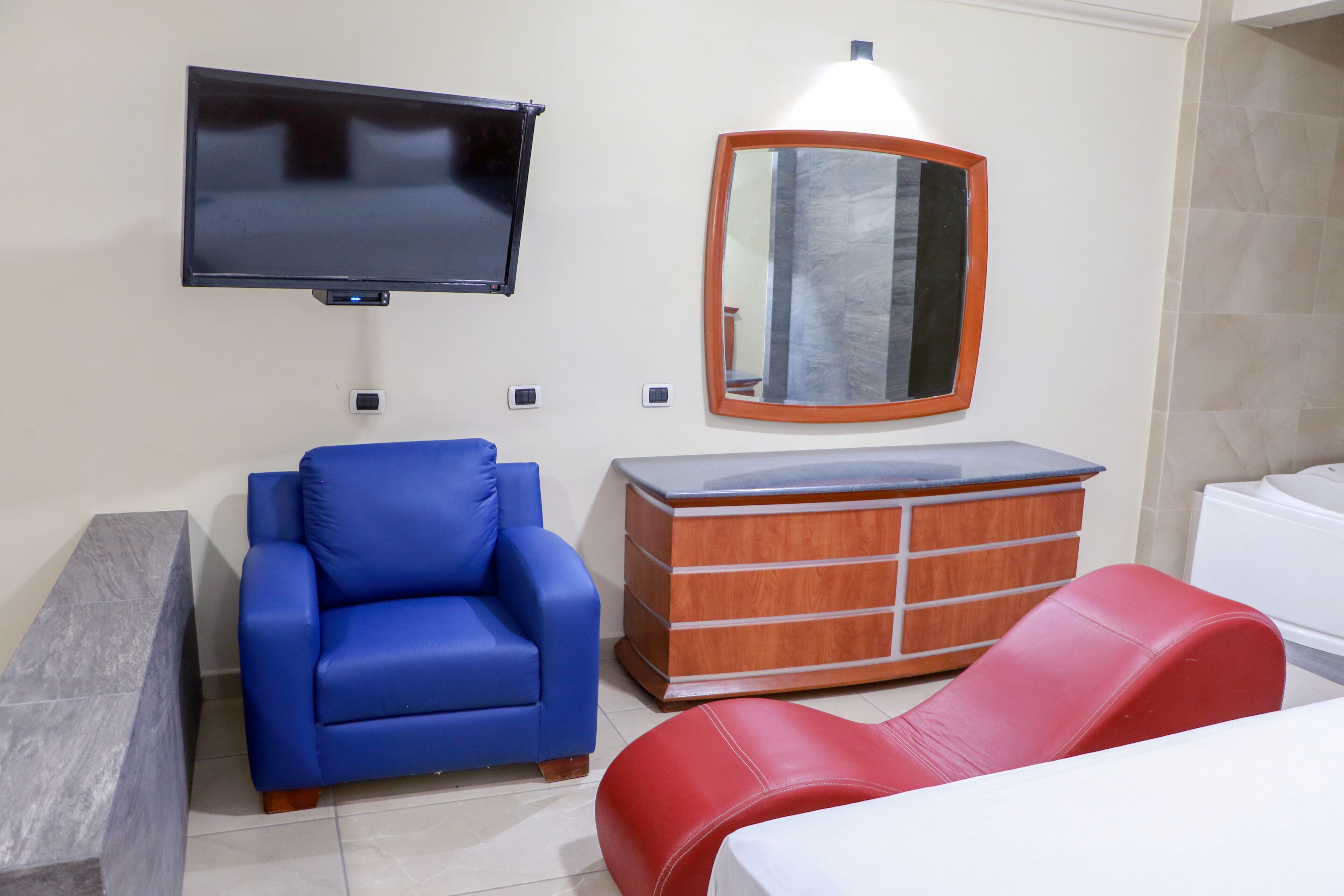 Une chambre d’hôtel équipée d’un téléviseur à écran plat, d’une commode en bois, d’un fauteuil bleu, d’une chaise rouge et d’un lit.