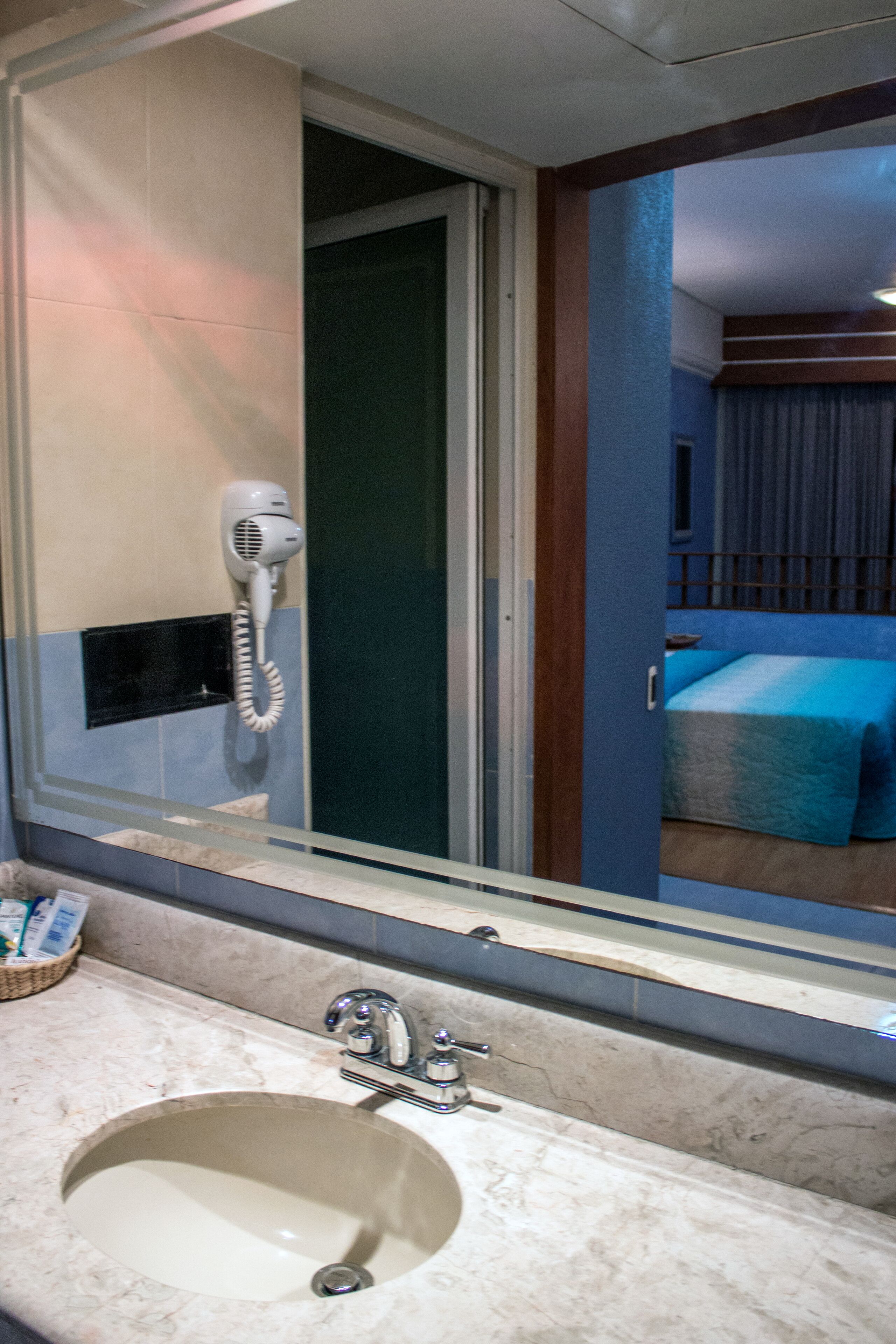 Et badeværelse med håndvask, hårtørrer og et spejl der viser et rum med seng og gardiner.