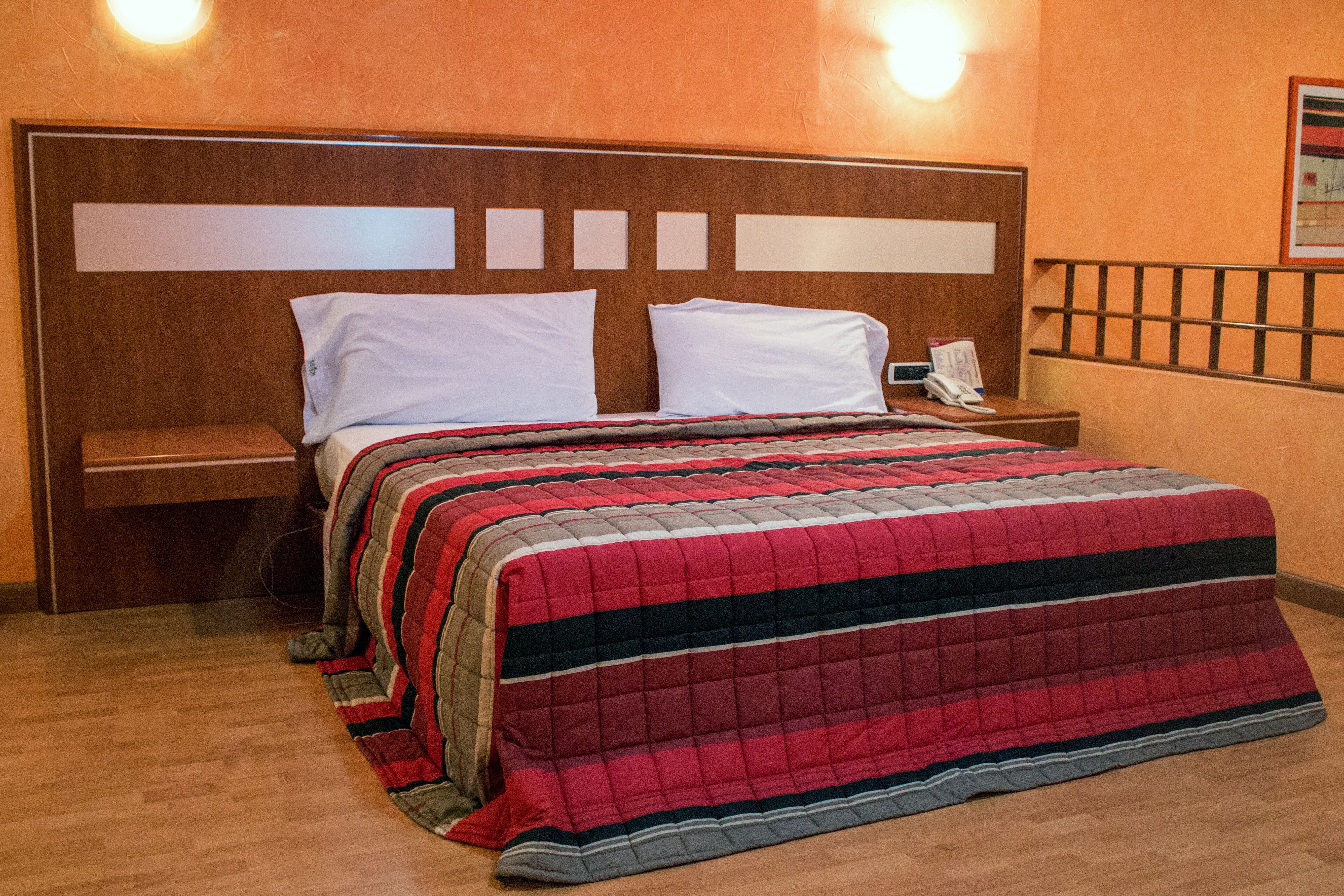 Une chambre d’hôtel avec une tête de lit en bois, deux oreillers blancs, une couvre-lit rouge et noire à rayures, une table de chevet en bois avec un téléphone et un plancher en bois.
