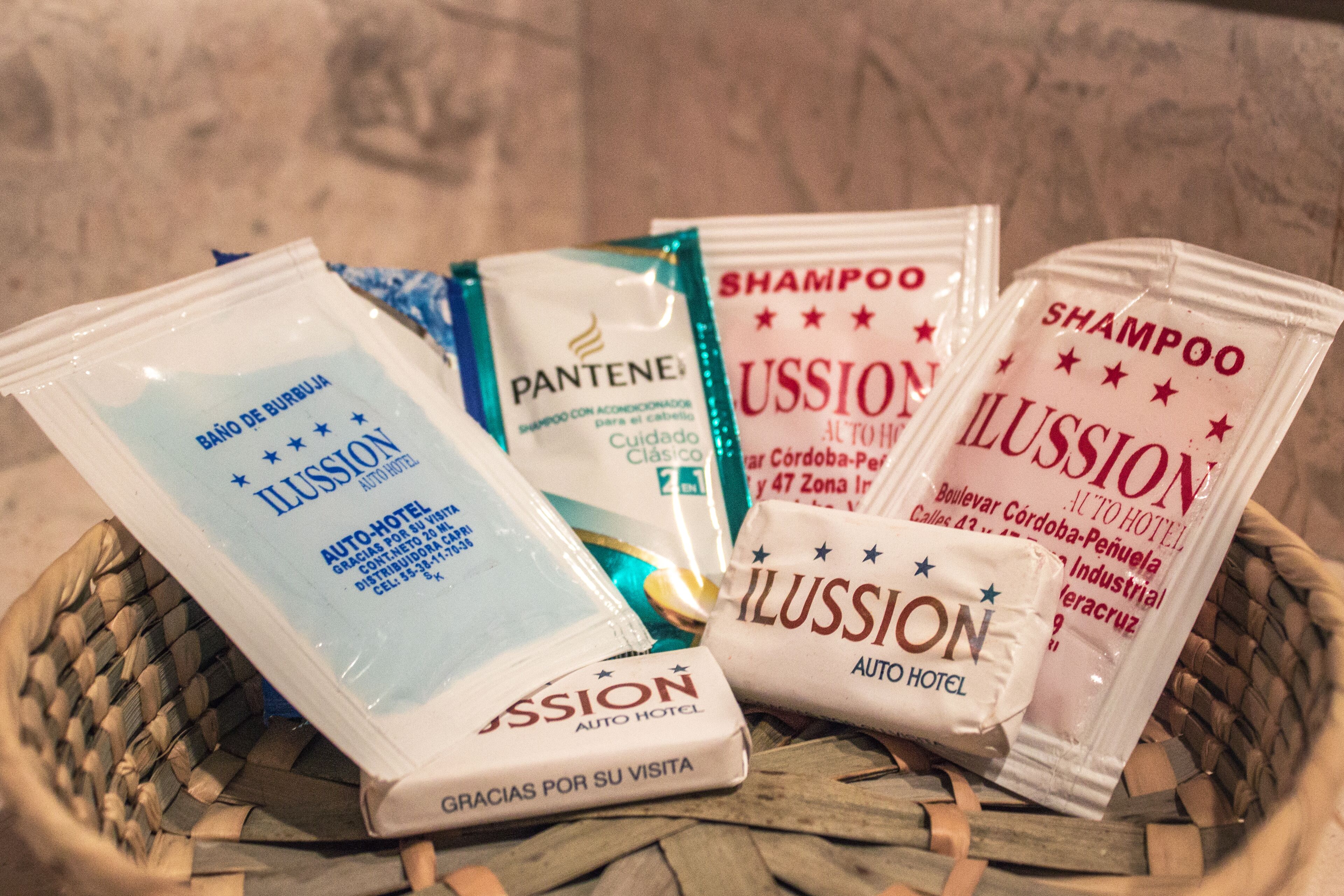 A basket containing various toiletries including a Pantene shampoo, a shampoo bar, and a toiletry bag from Illusion Auto Hotel.