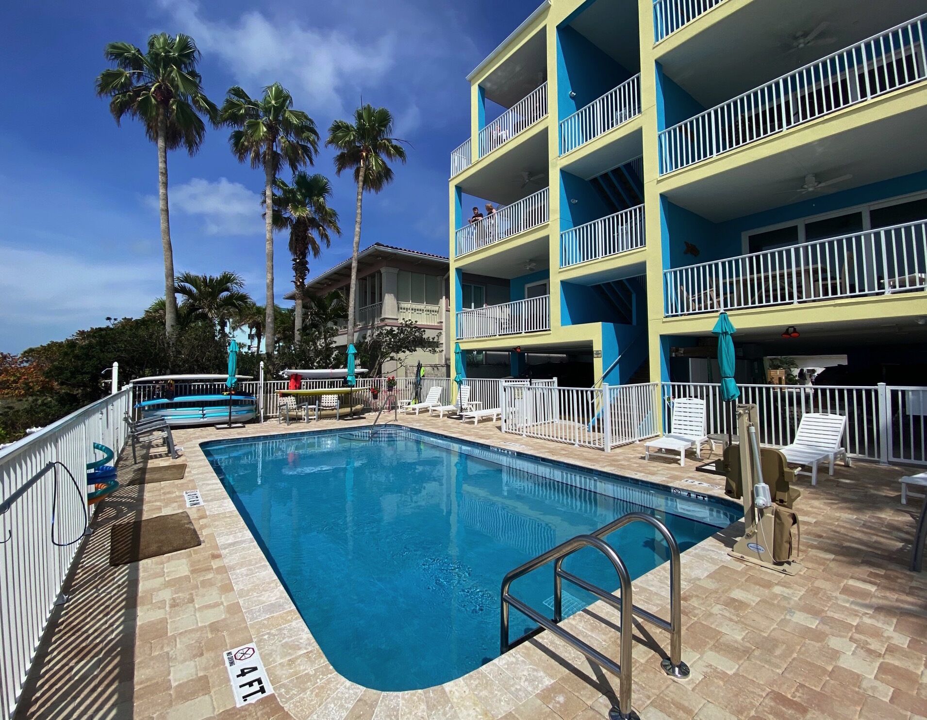 A swimming pool with a metal ladder, surrounded by a white fence and lounge chairs.