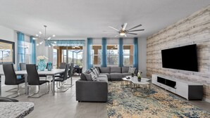 Un salon moderne avec un canapé d’angle gris, un coin repas avec une table et des chaises blanches, un téléviseur à écran plat fixé à un mur en bois et un ventilateur de plafond.