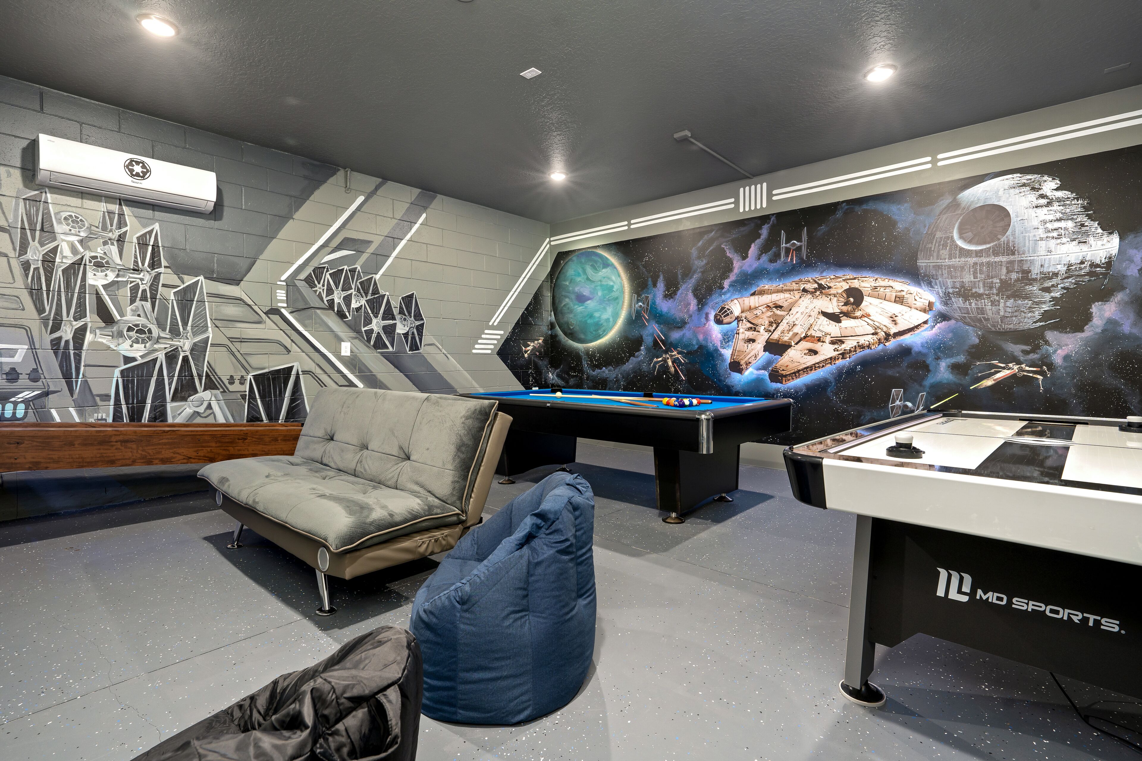 A pool table with a Star Wars mural, a sofa, and a bean bag in a room with a gray floor and white walls.