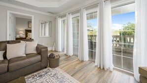 A spacious living room with a brown leather sofa, a patterned rug, and a large window with white curtains.