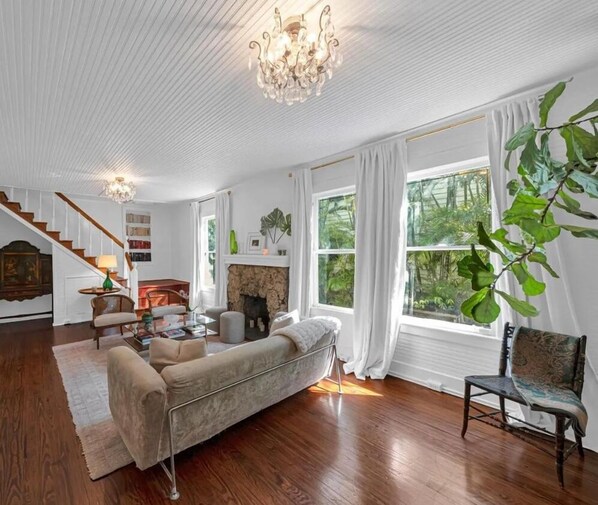A spacious living room with a fireplace, a chandelier, and a large window offering a view of greenery. - N.E. Coconut Grove pool home, family & pet friendly, large yard, & guest house. (Coconut Grove)
