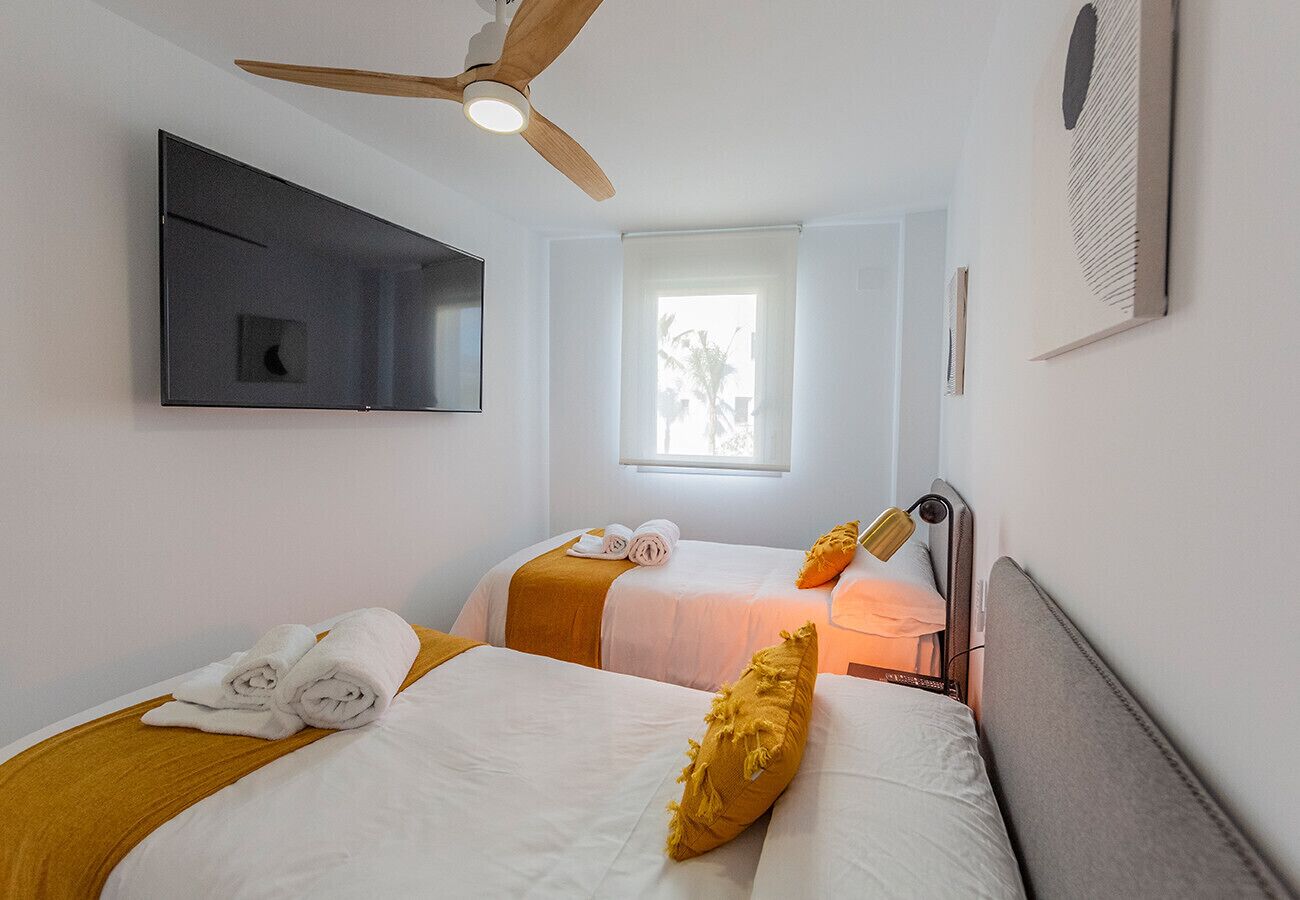 A hotel room with two beds, a ceiling fan, a TV mounted on the wall, and a window with a view of palm trees.