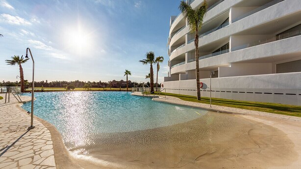 Una piscina con acqua cristallina circondata da un vialetto in pietra, palme e un edificio moderno con balconi.