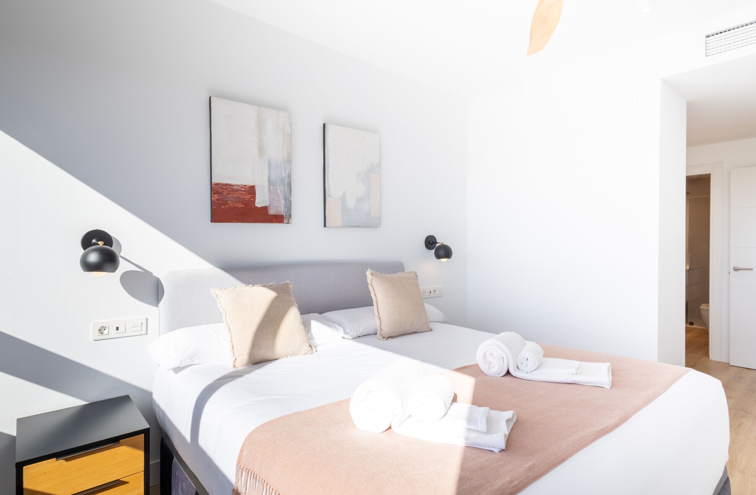 A modern hotel room with a neatly made bed, two wall-mounted artworks, and a bedside table with a lamp.