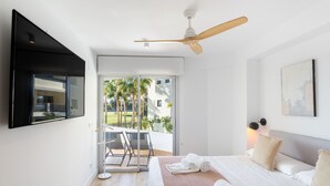 Une chambre d’hôtel moderne équipée d’un lit, d’un ventilateur de plafond, d’un téléviseur fixé au mur et d’un balcon offrant une vue sur les palmiers.