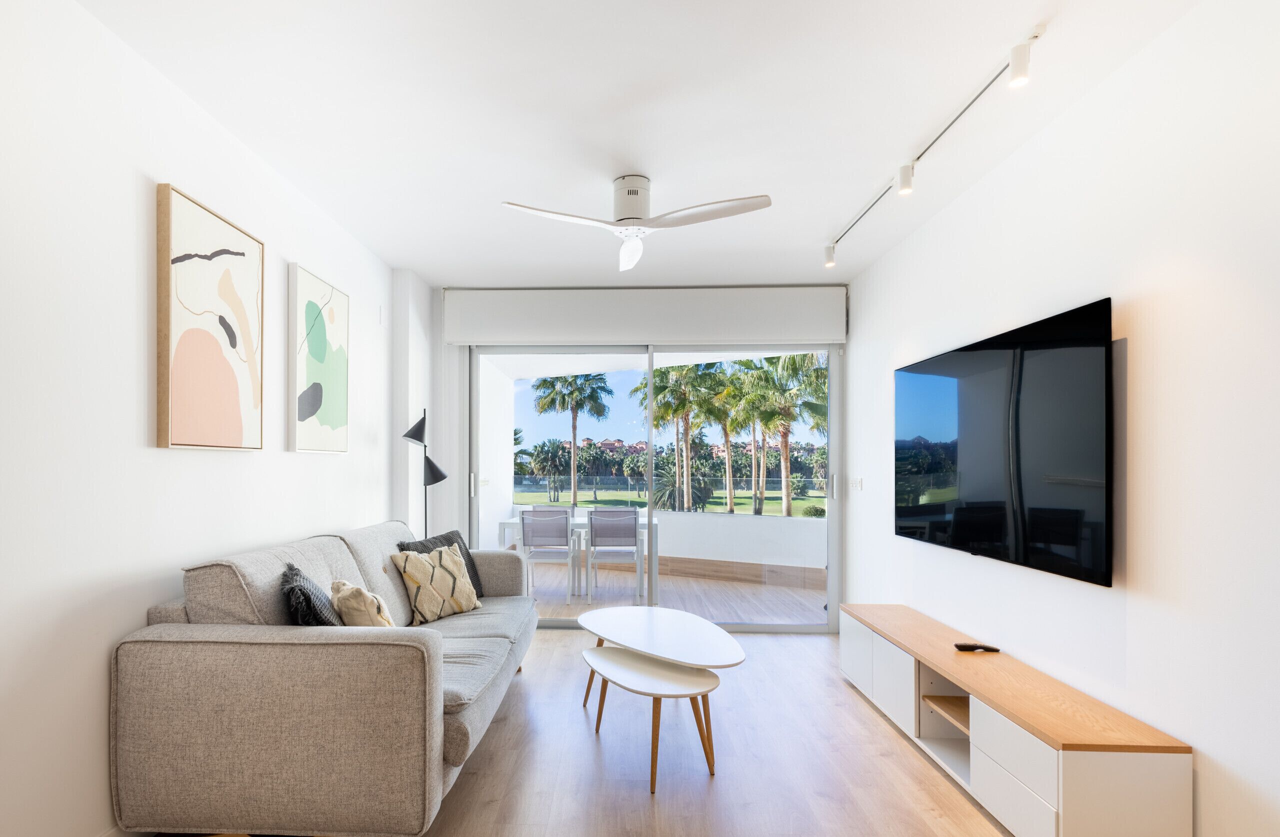 Una sala de estar moderna con sofá, mesa de centro y televisor. Hay dos cuadros enmarcados en la pared y un ventilador de techo.