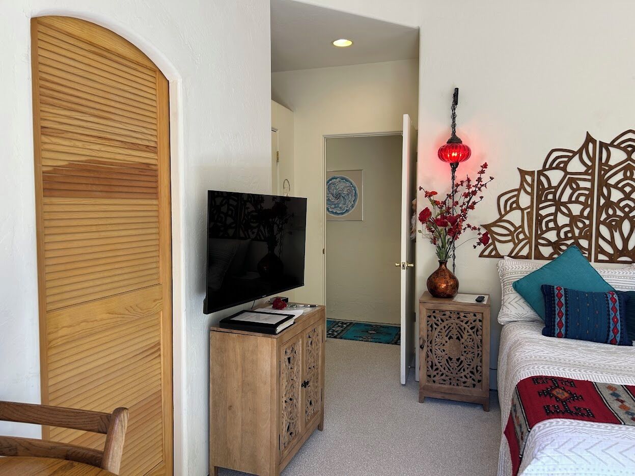 Een knus ingerichte kamer met een houten garderobe, een televisie op een houten kast, een decoratief wandobject en een bed met bedkussens met een patroon.