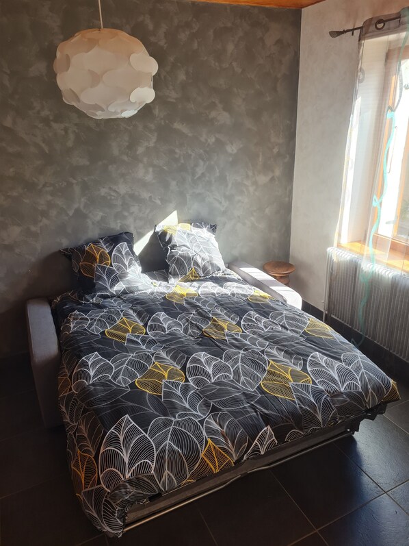 A bed with a patterned bedspread, a bedside table, and a hanging light fixture in a room with a gray wall and a window.