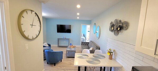 A modern living room with a large wall clock, a flat-screen TV, and a dining area with a white table and chairs. - Satellite Motel -  Shore to Please 109 (Treasure Island)