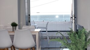 A modern room with a balcony overlooking the sea, featuring a dining area with white chairs and a small table, a potted plant, and a tiled floor with a decorative border.