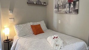 Un lit bien fait, avec une couette blanche et un oreiller orange, une serviette pliée posée sur le lit, une lampe de chevet et une étagère fixée au mur, ornée d’objets décoratifs.