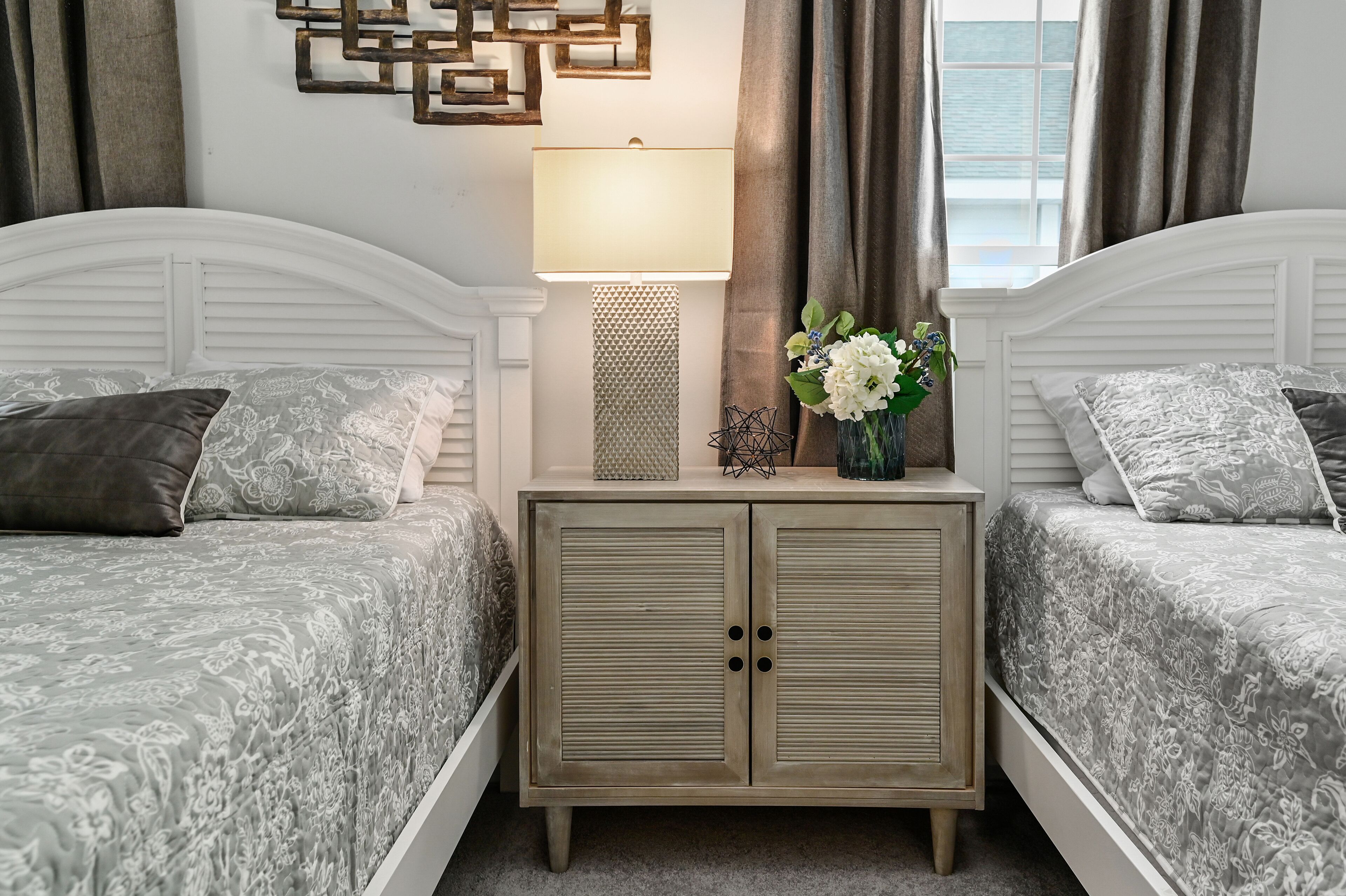 Een slaapkamer met twee bedden, een nachtkastje met lamp, een decoratief wandobject en een vaas met bloemen.