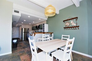 A dining area with a wooden table and white chairs, a hanging light fixture, and a wall decoration. - Aquarius - 207, Beachfront Condo located on the 2nd floor overlooking the beach & Gulf of Mexico. (South Padre Island)