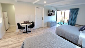 A hotel room with two beds, a dining area with a table and chairs, a television on the wall, and a sliding glass door to the outside. - Satellite Motel - Shore Thing 111 (Treasure Island)