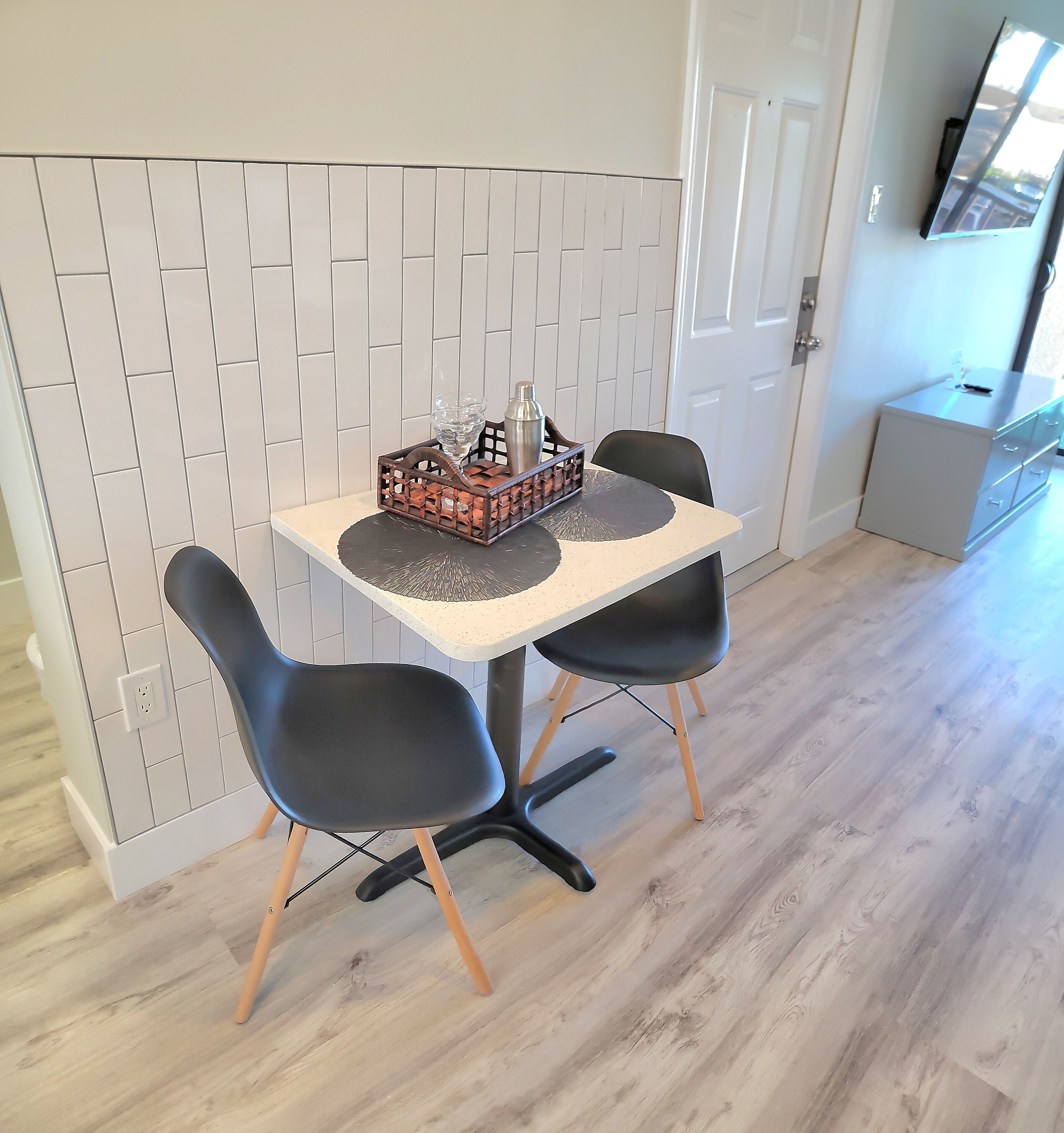 A dining area with a table and two chairs, a decorative tray with a glass and a bottle, and a white tiled wall with a door in the background.