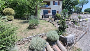 A two-story stone house with a balcony, surrounded by a garden with various plants and a stone pathway.